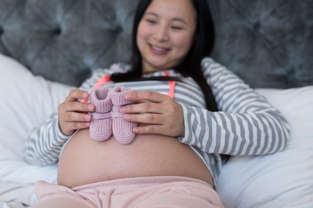 This image shows a pregnant woman holding baby socks on her belly while sitting in a bedroom. She is smiling and appears happy and excited about the upcoming arrival of her baby. This image can be used for maternity blogs, parenting websites, prenatal care articles, or advertisements for baby products.