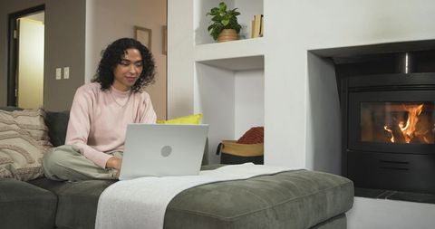 Nonbinary person working on laptop in cozy living room by fireplace, relaxing on gray sofa