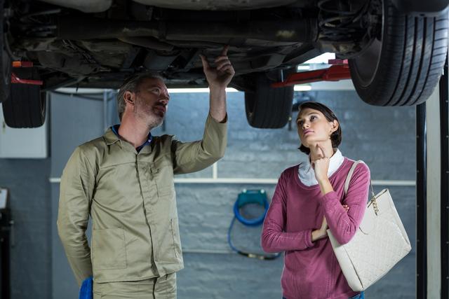 Mechanic in uniform showing car issue to a concerned female customer in an auto repair shop. Useful for illustrating automotive services, customer service in the automotive industry, car maintenance, and repair shop environments.