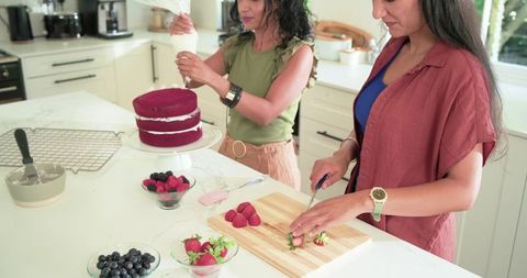 Two diverse women decorating red velvet three-layer cake in modern kitchen, slicing strawberries