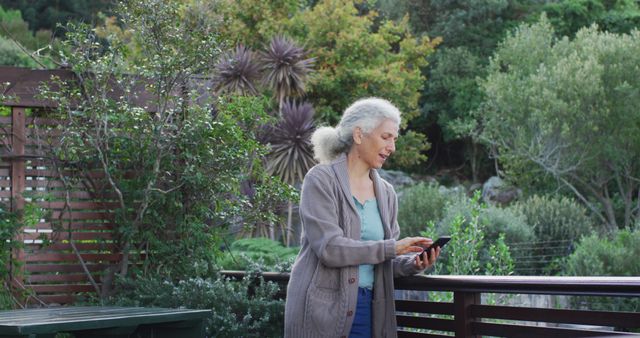 An elderly woman interacts with her smartphone on a balcony surrounded by a lush garden. This scene highlights the harmony between technology and nature, making it ideal for themes of active aging, digital literacy among seniors, relaxation, and enjoying nature. Useful for campaigns on healthy lifestyle, outdoor relaxation, or tech use among older adults.