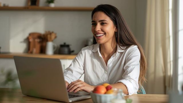 Smiling woman working remotely on laptop in sunlit minimalist kitchen, casual white shirt