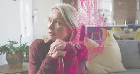 Contemplative mature woman sitting with cane looking in sunlit living room with pink overlay
