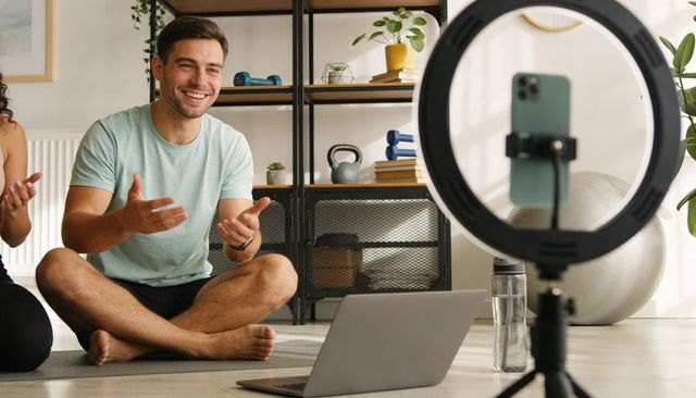 Smiling man hosting livestream workout from home using ring light, laptop and yoga mat