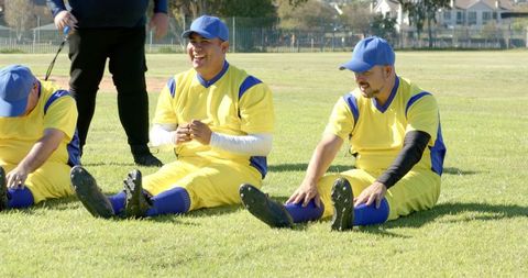 Diverse male baseball teammates stretching and laughing on grass field in yellow uniforms
