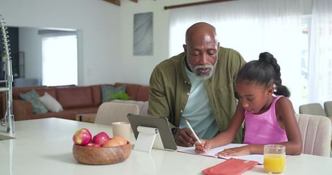 Grandfather tutoring granddaughter at kitchen island using tablet for homework and learning