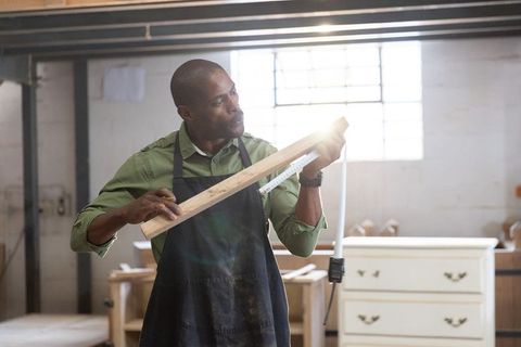 African american carpenter measuring wooden plank in sunlit workshop with tape measure