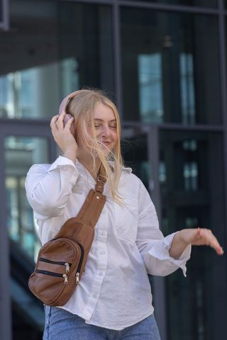 Young woman enjoying music with pink headphones and brown leather sling bag at urban glass facade