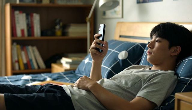 Teenage asian male lying on bed using smartphone in bedroom with blue duvet, casual grey tee