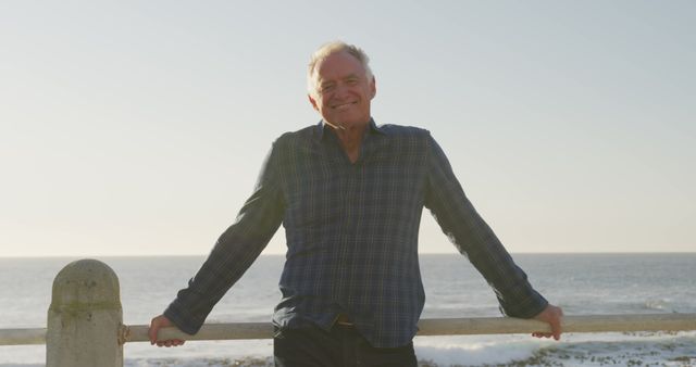 Elderly man leaning on railing by ocean, smiling happily in the sunlight. Ideal for ads about retirement, healthy lifestyle for seniors, positivity in old age, and outdoor activities for retirees.