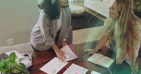 Two women leaning over charts and notebook, collaborating on business planning at home