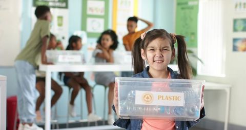 Elementary girl holding plastic recycling bin in classroom teaching waste sorting