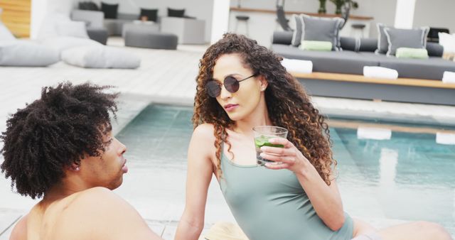 A couple is relaxing poolside, with the woman holding a cocktail and wearing sunglasses in a modern outdoor space. They both appear to be enjoying a leisurely summer day. This can be used for lifestyle blogs, vacation promotions, swimwear advertisements, or articles on summer leisure activities.
