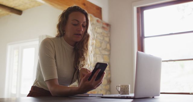 Young woman wearing casual clothing using smartphone while working on laptop in modern home office with large window and bright lighting. Ideal for concepts of remote work, technology use, communication, multitasking, and modern lifestyle.