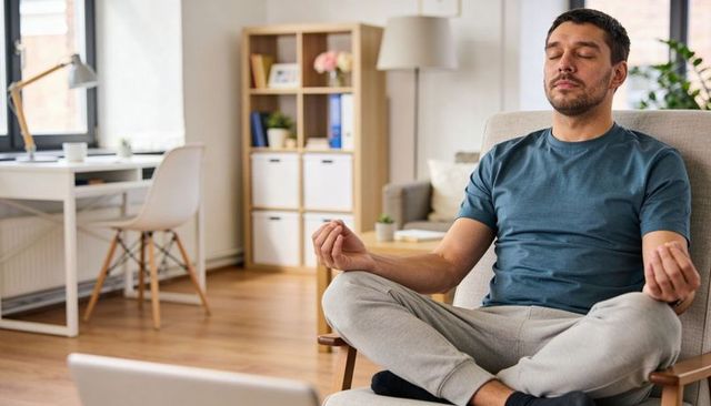 Meditating man sitting cross-legged in modern home office chair with laptop for wellness