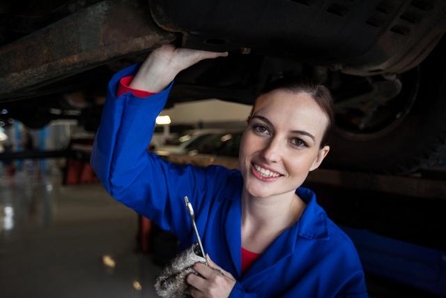 Female mechanic smiling while servicing a car in a garage. She is wearing a blue uniform and holding a wrench, working on the undercarriage of a vehicle. This image can be used for promoting automotive services, illustrating articles about women in skilled trades, or showcasing professional mechanics at work.