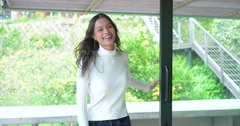 Smiling woman stepping onto balcony through sliding door wearing white sweater, jeans