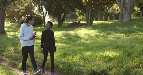 Young adults walking and talking on sunlit park trail while tracking fitness on phone