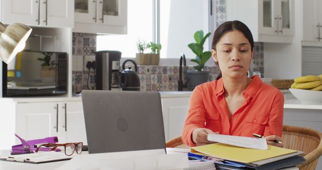 Young woman reviewing papers and finding financial documents while working at a laptop in a modern kitchen home office setup. Use for concepts related to remote work, financial planning, home office productivity, and modern lifestyles.