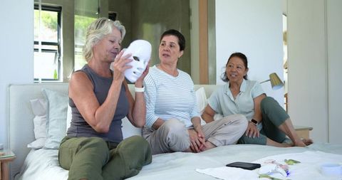 Mature friends exploring skincare and self-care devices while relaxing on bed together
