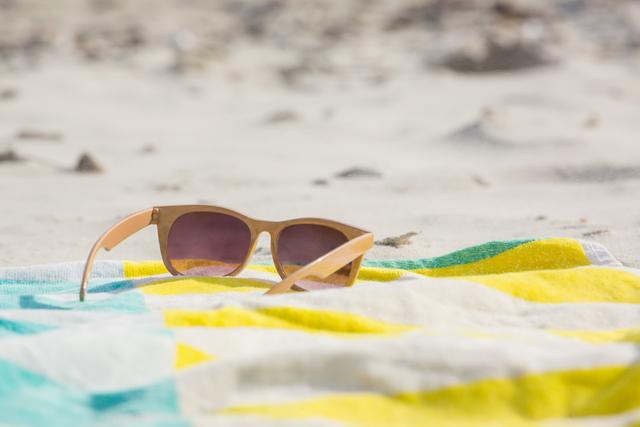 Sunglasses resting on a colorful beach blanket at a sandy beach. Ideal for use in travel blogs, summer vacation promotions, eyewear advertisements, and leisure activity websites.