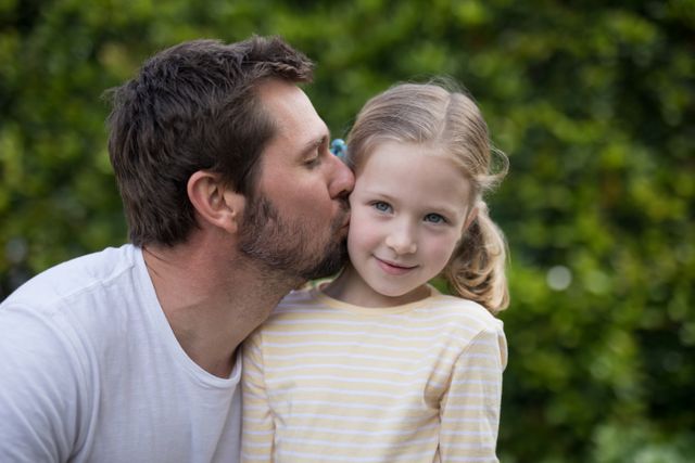 Father showing affection by kissing his young daughter on the cheek in a park. The girl is smiling and wearing a striped shirt. Ideal for use in family-oriented advertisements, parenting blogs, and articles about father-daughter relationships.