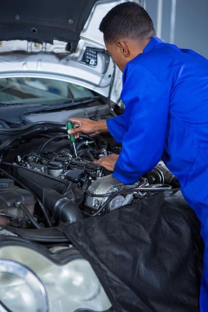 Mechanic in blue uniform working on car engine in repair garage. Ideal for content related to car maintenance, automotive industry, professional repair services, and technical expertise.