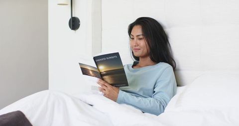 Asian woman reading in bed wearing blue pajama top relaxing under wall sconce light