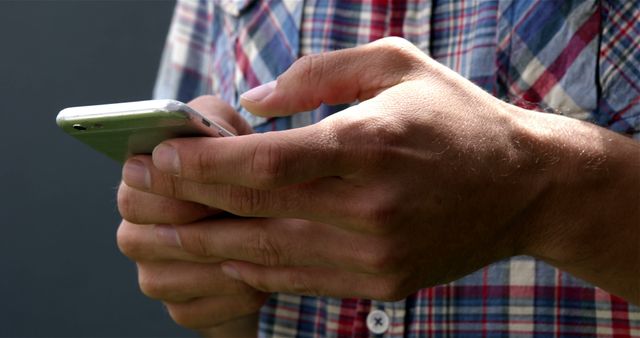 Close-up photo of person's hands texting on a smartphone while wearing a plaid shirt outdoors. Useful for illustrating themes like communication, technology, on-the-go lifestyle, and modern mobile text usage.