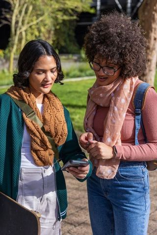 Multicultural young women sharing smartphone and skateboard in sunny urban park