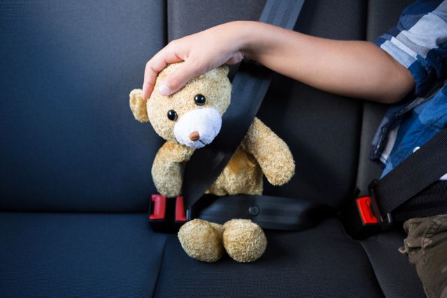 Child securing teddy bear with seatbelt in car back seat. Ideal for use in articles or advertisements about child safety, travel tips, family road trips, and transportation safety. Can also be used in parenting blogs, educational materials, and toy promotions.