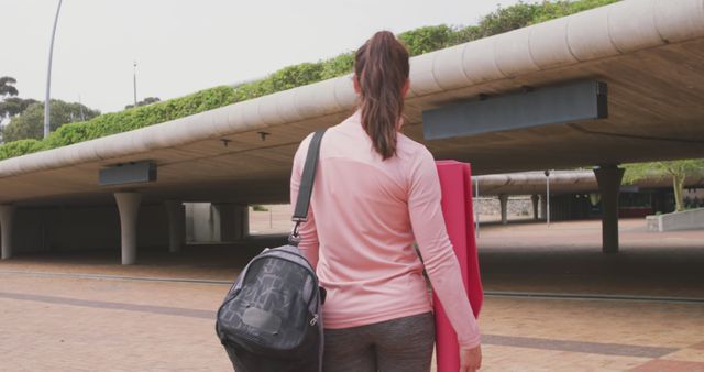 Woman dressed in sportswear carrying a red yoga mat and a gym bag, heading towards an outdoor area. Useful for topics relating to fitness, healthy lifestyle, outdoor exercises, or yoga practice. Ideal for promotions, blog posts, or articles about staying active and healthy.