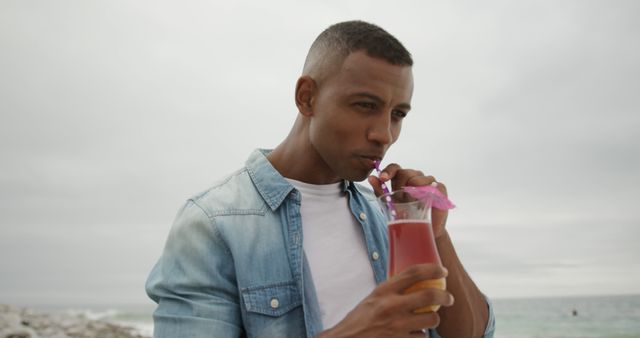 Happy african american man wearing denim jacket drinking cocktail on beach. Lifestyle, summer, vacation and leisure, unaltered.