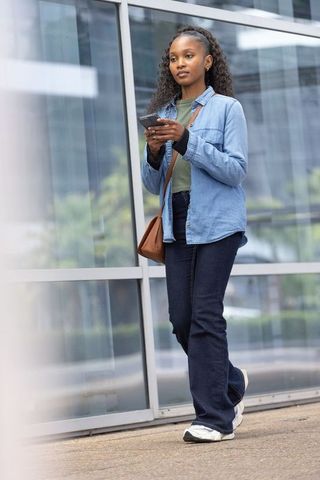 African american woman walking and texting on smartphone by glass facade, urban commuter