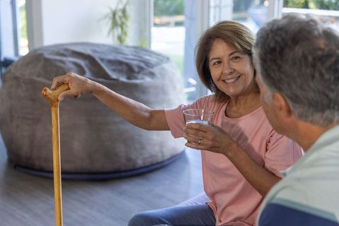 Senior woman offering glass of water while holding wooden cane, smiling with companion