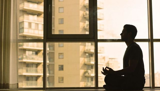 Urban mindfulness: man meditating cross-legged by high-rise window silhouette