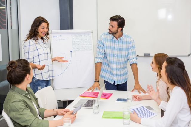 Team of executives discussing business strategy around a table in an office. One executive is presenting data on a flip chart while others listen and engage. Ideal for illustrating teamwork, corporate meetings, business planning, and professional collaboration in a modern office environment.