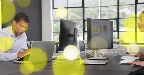 Young professional writing notes at laptop in modern open-plan office with data charts