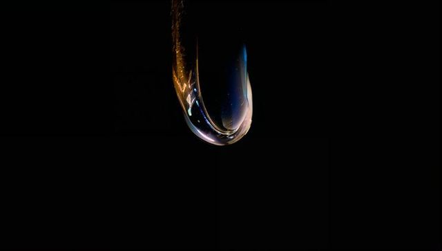 Iridescent teardrop droplet suspended on thin filament reflecting colorful refractions and glow