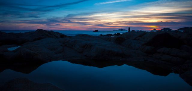 Scene captures serene rocky shore at twilight, lone silhouette on rocks, reflections in calm water, vibrant twilight sky. Ideal for peaceful, tranquil themes, nature, travel promotions, backgrounds, posters.