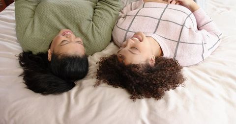 Two Female Friends Lying on Bed Laughing in Cozy Knit Sweaters Relaxing Together