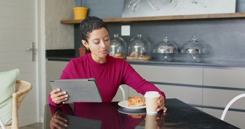 Working on tablet and reaching for coffee, woman enjoying pastry at modern kitchen table