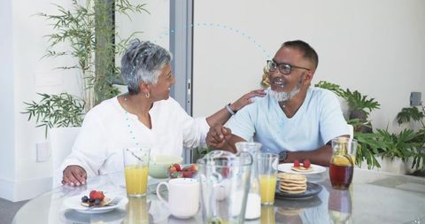 Senior couple holding hands and sharing sunlit breakfast, smiling over pancakes at glass table