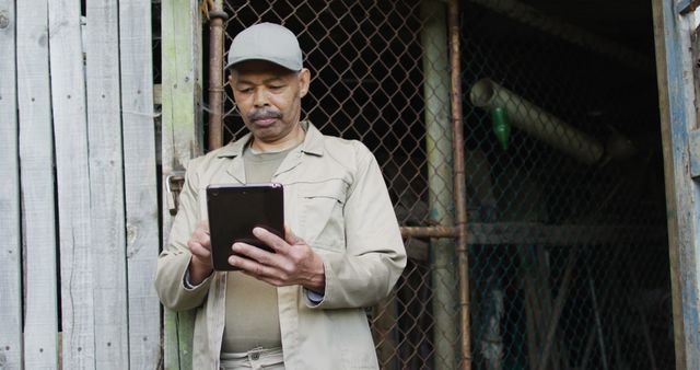 Elderly man is using a tablet while standing outside a rustic shed in a rural area. He is dressed in casual work clothes, including a cap and jacket. This image can be used to represent themes of technology adoption among older adults, rural life, and digital connectivity in remote areas.