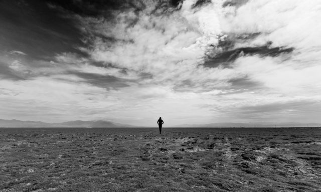 Solitary figure standing in middle of vast desert, under expansive cloudy sky. Monochrome enhances impression of solitude and isolation. Ideal for themes of solitude, introspection, nature's expanse, and minimalist art. Suitable for backgrounds, visual storytelling or metaphors for loneliness.