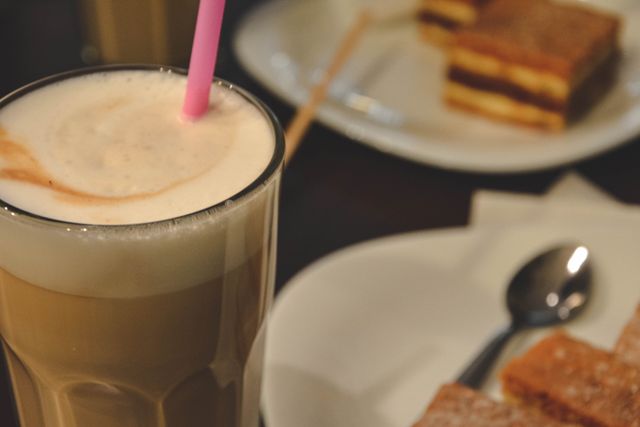 Close-up view of creamy latte with pink straw alongside pastries on plates in cafe. Ideal for coffee shop promotions, food and beverage blogs, and café menu designs.