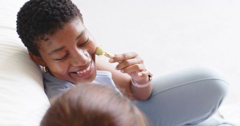 African american woman using jade facial roller while relaxing on bed with friend