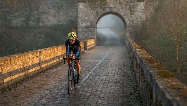 Female cyclist pedaling across misty stone bridge at golden hour with yellow helmet