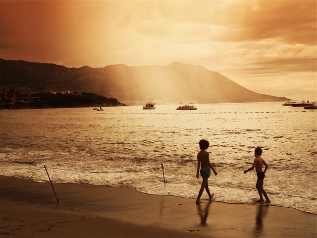 Two children enjoy a tranquil walk along the beach at sunset. Silhouetted by the golden tones of the setting sun, they stroll on the wet sand with the calm sea waves hitting their feet. In the background, gentle waves lap over boats anchored near the shore, while the sun sets behind picturesque mountains. Ideal for use in travel brochures, summer vacation advertisements, or family recreation themes to invoke a sense of peace and leisure.