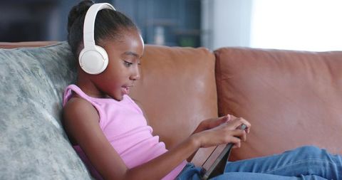African american girl wearing headphones using tablet on leather couch for remote learning